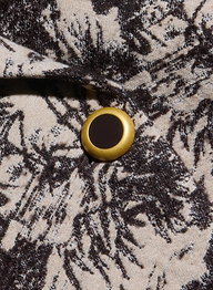 A close-up of a gold and black button on beige fabric adorned with the Kaisley Blazer’s dark, intricate leafy pattern, featuring its antique-inspired print.