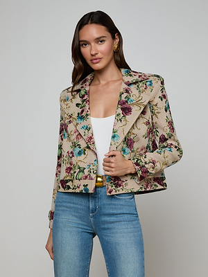 A woman with long brown hair stands confidently against a plain backdrop, wearing blue jeans, a white top, and the Xoey Cropped Trench Coat featuring chic pink, purple, and green floral tapestry accents.