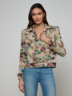 A woman with long brown hair wears the Xoey Cropped Trench Coat—a beige floral tapestry jacket with pink, green, and blue patterns—paired with light blue jeans, standing against a plain light gray background.