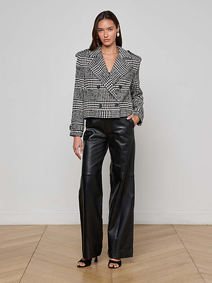 A woman stands against a light gray wall, wearing the Xoey Cropped Trench Coat with black wide-leg leather pants and black open-toe heels. Her hair is down and one hand is in her pocket.