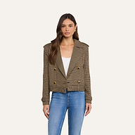 Una donna indossa il trench corto Xoey, un blazer marrone a quadri a doppio petto con bottoni dorati, sopra un top bianco e dei jeans blu; è in piedi davanti a uno sfondo beige a tinta unita e guarda verso la fotocamera.