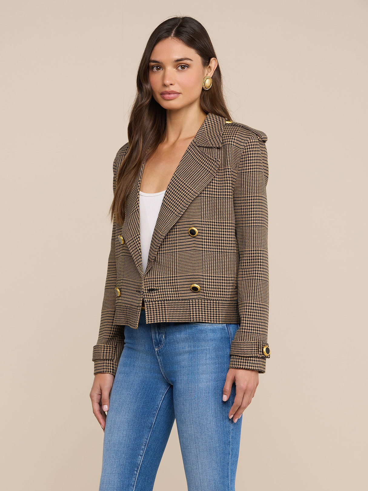 Una donna dai lunghi capelli castani indossa il trench corto Xoey sopra un top bianco, jeans blu e orecchini dorati, in piedi davanti a uno sfondo beige a tinta unita.