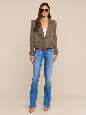 A woman wearing sunglasses, the Xoey Cropped Trench Coat over a white top, blue bootcut jeans, and tan heels stands against a plain beige background.