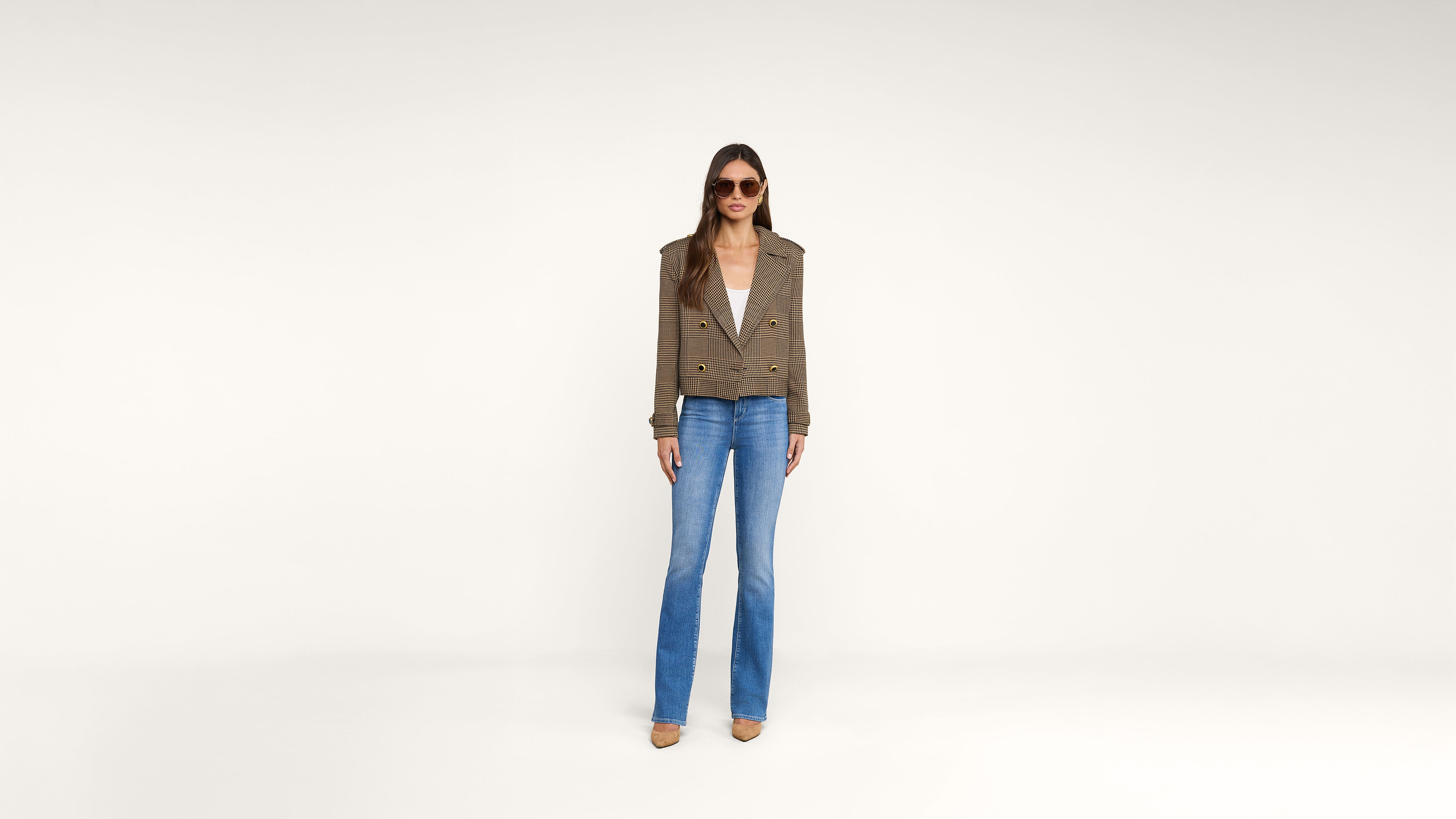 A woman wearing sunglasses, a brown plaid blazer, white top, blue jeans, and brown shoes stands against a plain white background.