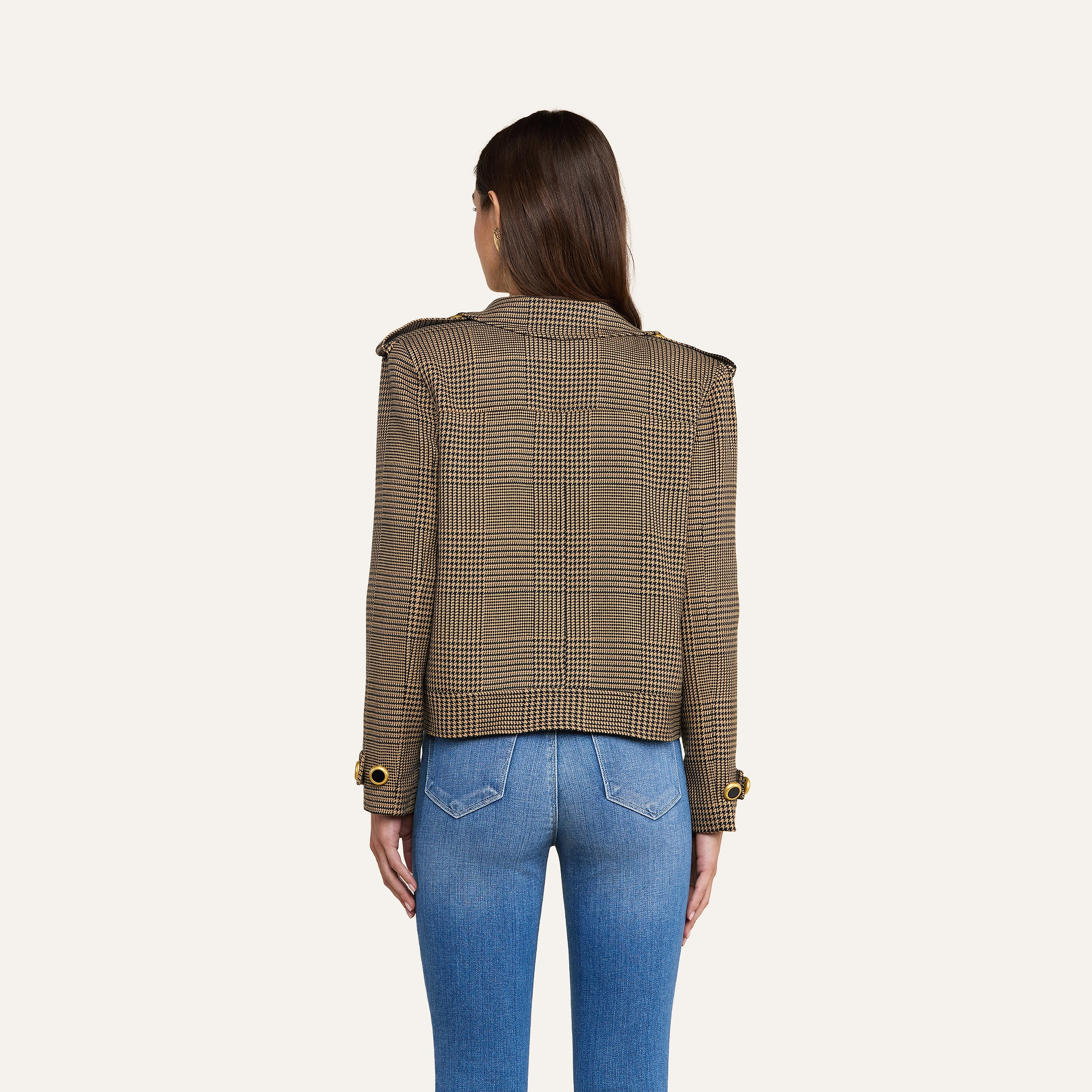 Si vede una donna di spalle che indossa il trench corto Xoey — un blazer a quadri marroni con polsini abbottonati — abbinato a jeans azzurri, in piedi davanti a uno sfondo beige a tinta unita.