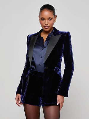A woman poses confidently in power dressing style, wearing the L'AGENCE Sheila Velvet Longline Blazer in dark blue with black satin lapels, paired with a matching shirt and velvet shorts over sheer black tights.