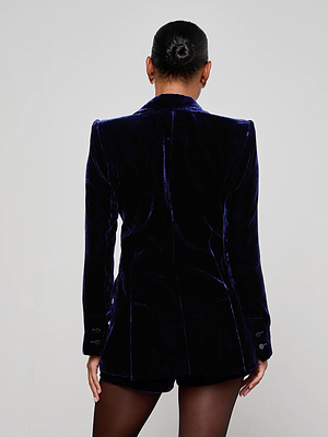 A person with dark hair in a neat bun is seen from the back, wearing the Sheila Velvet Longline Blazer by L'AGENCE with matching shorts and black tights, set against a plain light gray background.