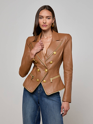 A woman with long brown hair models the L'AGENCE Ottilie Leather Blazer—fitted Italian leather with gold buttons and zippers—paired with blue jeans, posing against a plain light gray background.