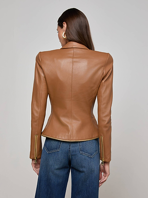 A woman faces away, wearing the L'AGENCE Ottilie Leather Blazer with zippered sleeves and blue jeans. Her straight brown hair is down, gold hoop earrings add polish, and she stands before a plain light background.