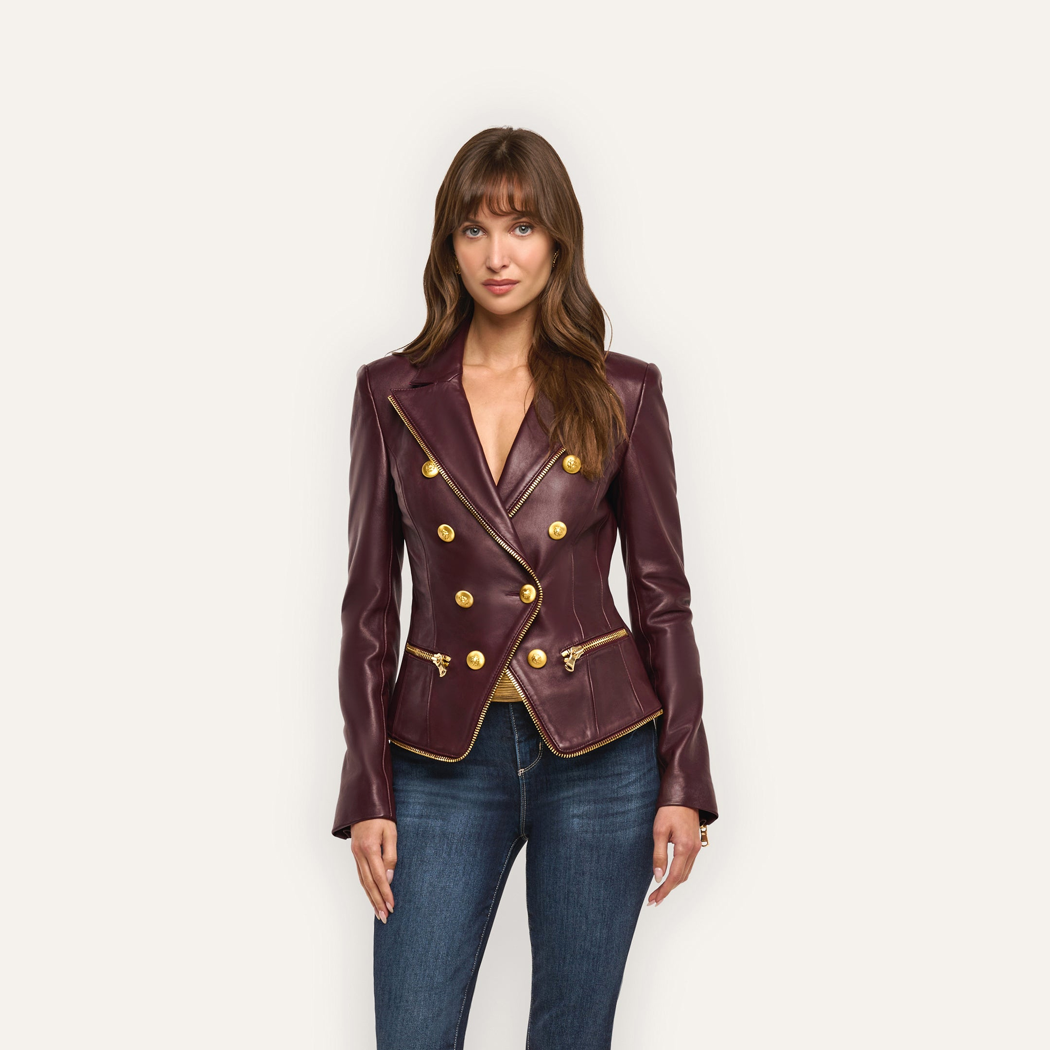 A woman wears the Ottilie Leather Blazer in deep burgundy, featuring a double-breasted gold button closure and gold zippered pockets for a sharp, hourglass power-dressing silhouette.