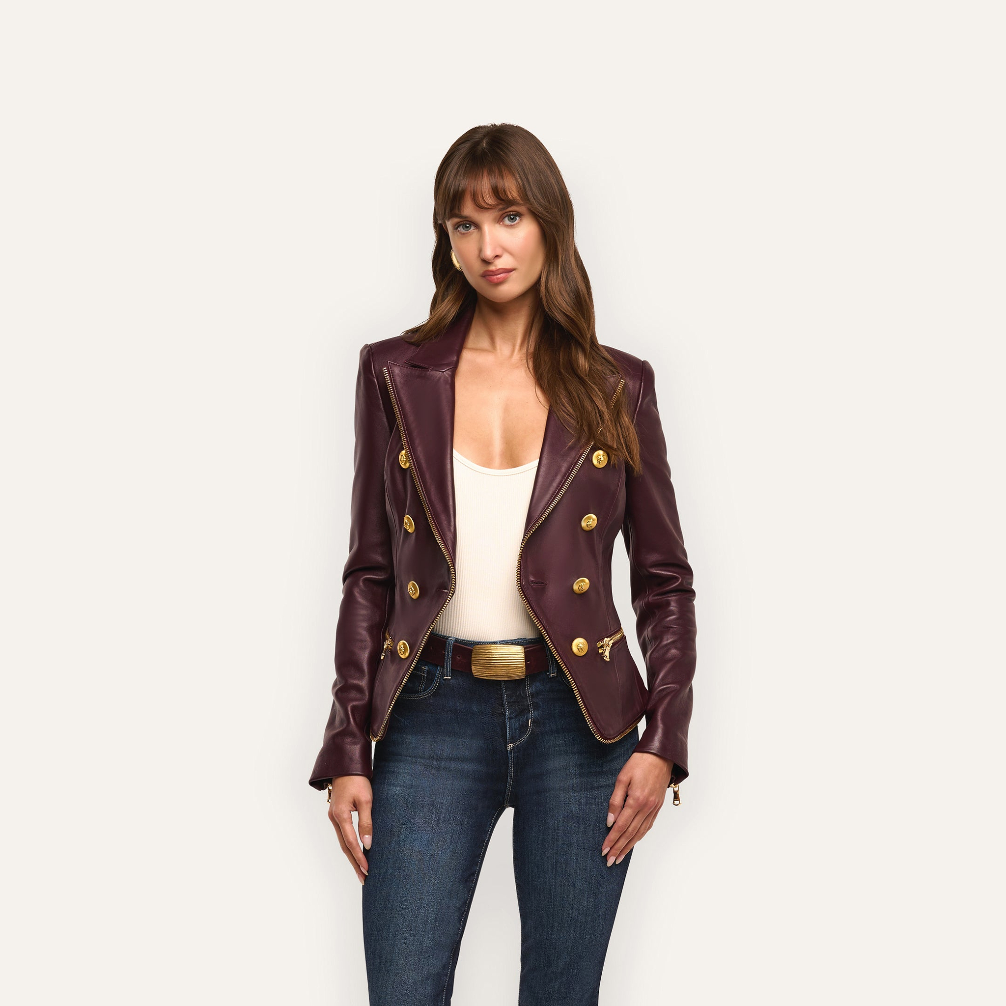 A woman in an hourglass silhouette wears an Ottilie Leather Blazer in dark burgundy with gold buttons over a white top and dark-wash jeans.