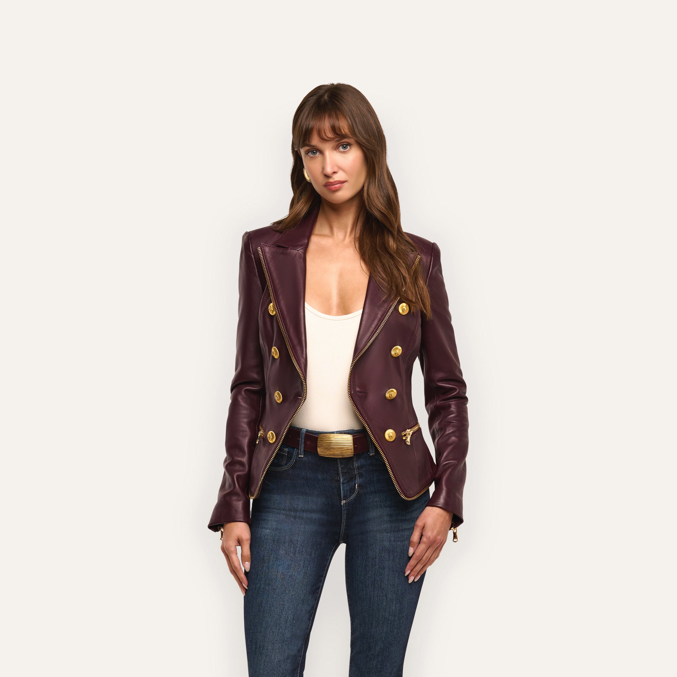 A woman in an hourglass silhouette wears an Ottilie Leather Blazer in dark burgundy with gold buttons over a white top and dark-wash jeans.