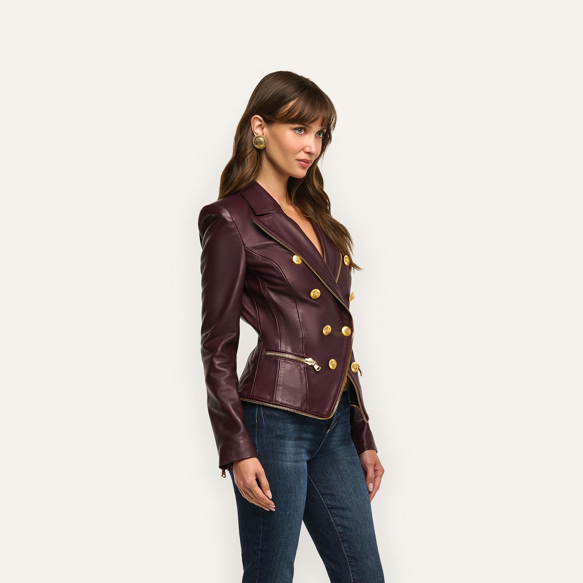 A woman in an hourglass silhouette wears an Ottilie Leather Blazer in dark burgundy with gold buttons and zipper accents, paired with blue jeans for a look of power dressing.
