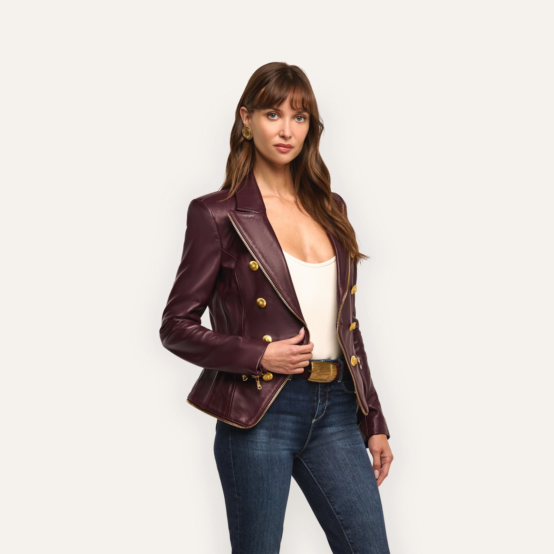 A woman in an hourglass silhouette wears an Ottilie Leather Blazer in burgundy with gold buttons over a white top and blue jeans, embracing power dressing.