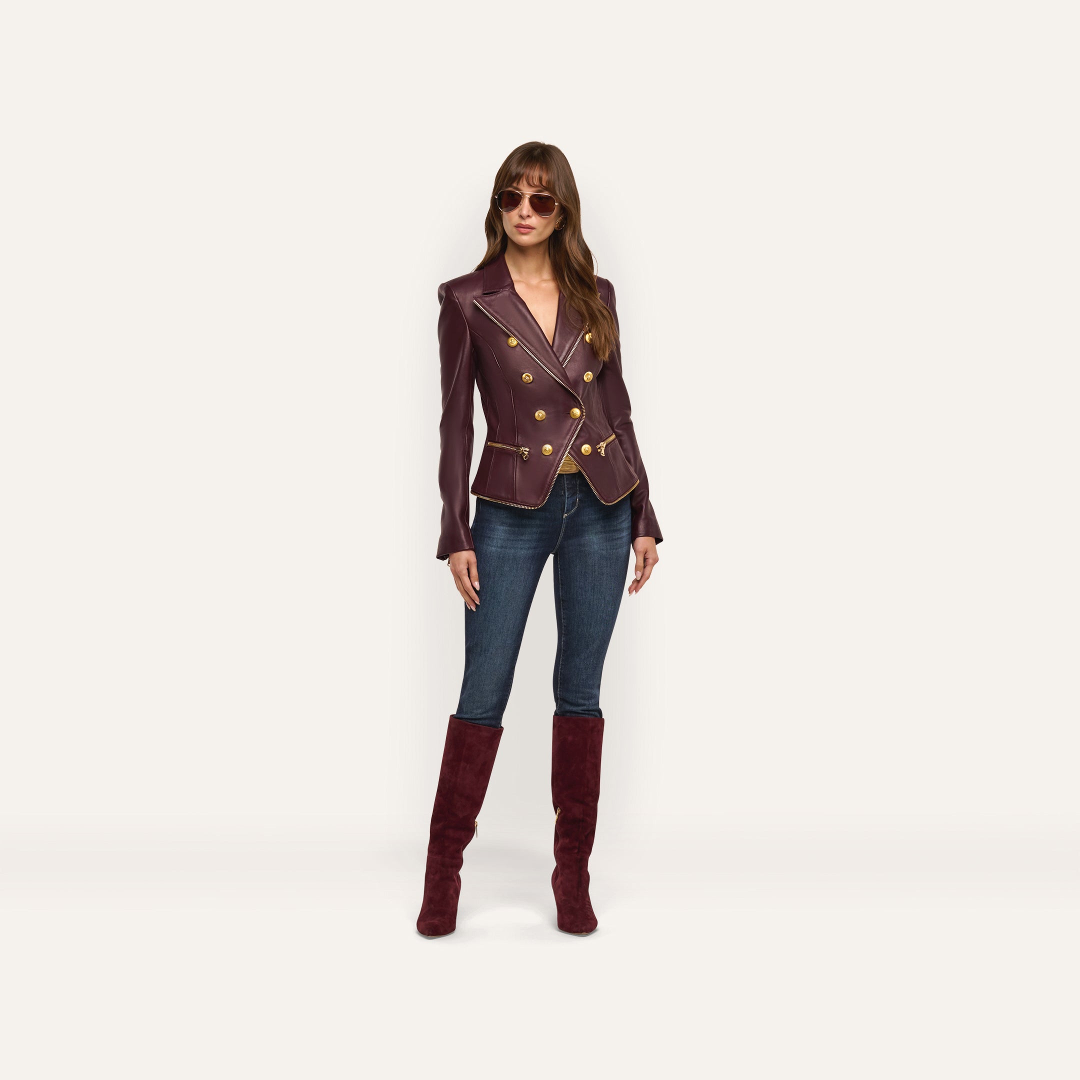 A woman creates an hourglass silhouette in a burgundy double-breasted Ottilie Leather Blazer with gold buttons, dark-wash jeans, and matching knee-high suede boots for a look of power dressing.