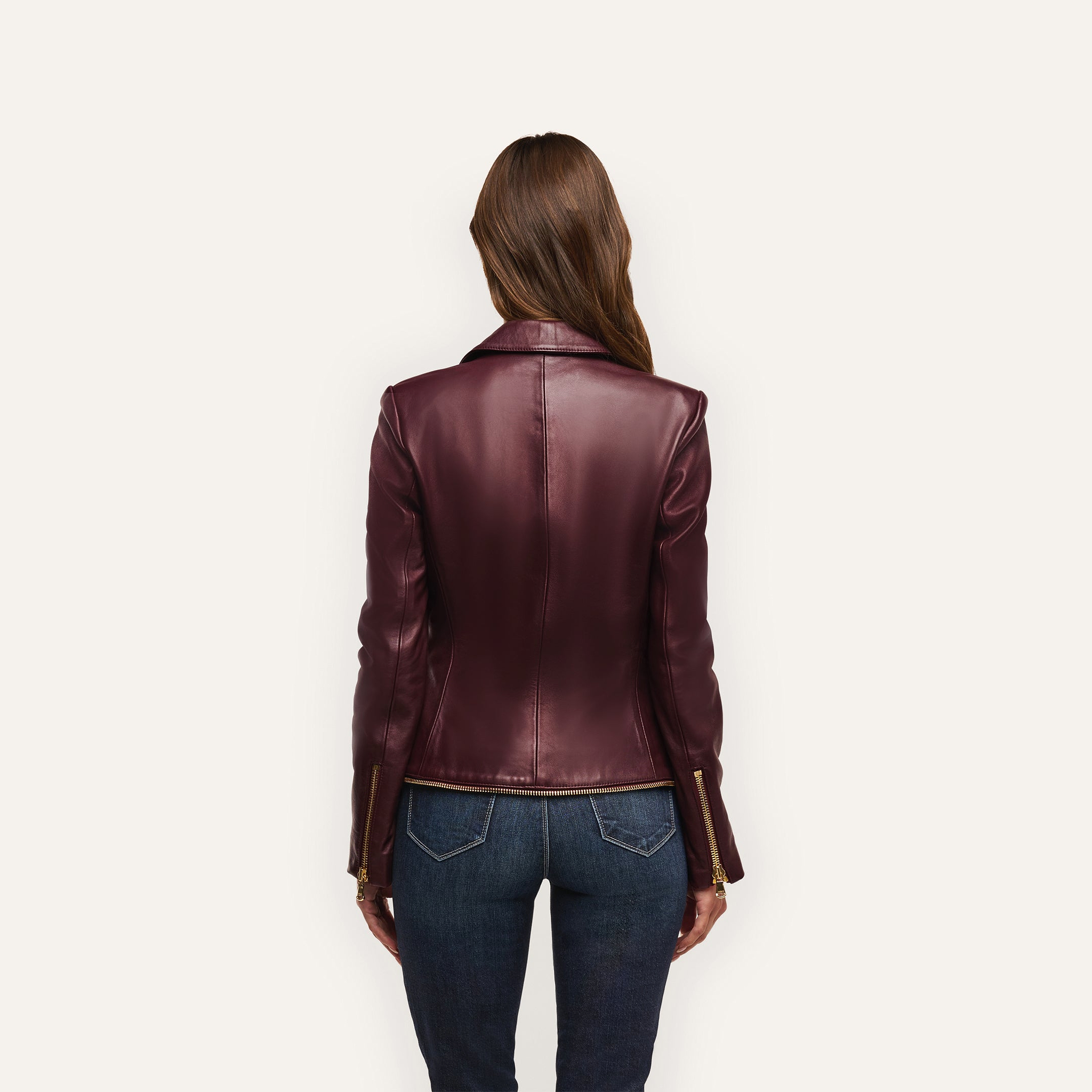 A person seen from behind wearing an Ottilie Leather Blazer in burgundy with gold sleeve zippers and blue jeans, the outfit creating a sharp power dressing look with an hourglass silhouette.