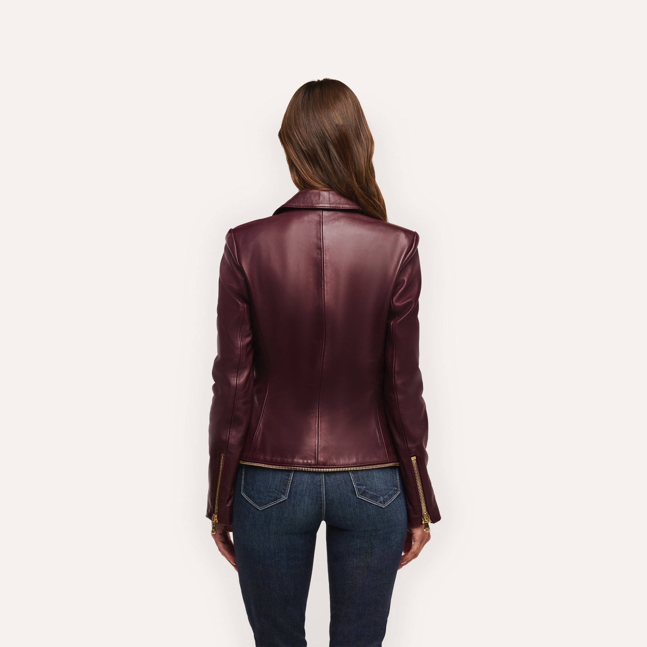 A person seen from behind wearing an Ottilie Leather Blazer in burgundy with gold sleeve zippers and blue jeans, the outfit creating a sharp power dressing look with an hourglass silhouette.