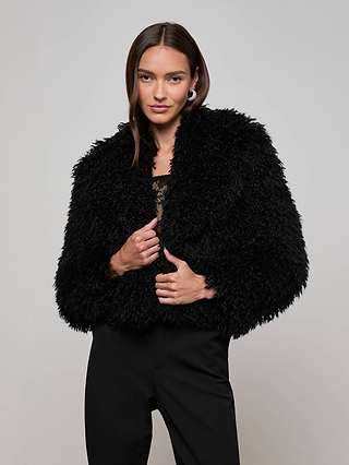 A woman with long brown hair wears the Aura Cropped Faux Fur Jacket by L'AGENCE over a lace top and black pants, standing against a plain light background and looking confidently at the camera.