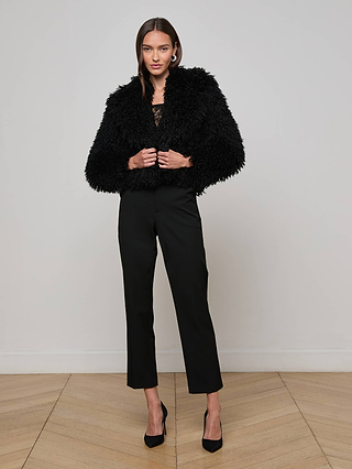 A woman stands indoors against a plain wall wearing the L’AGENCE Aura Cropped Faux Fur Jacket, black cropped pants, and high heels. She has straight brown hair and looks at the camera with her hands on her jacket.