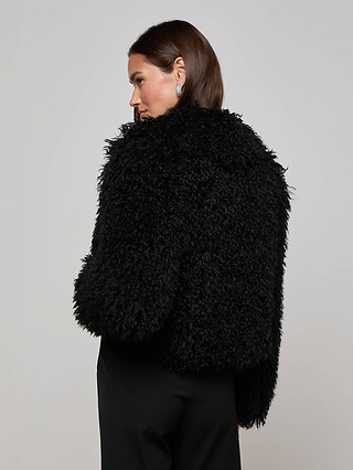 A woman with straight brown hair, facing away, wears the L'AGENCE Aura Cropped Faux Fur Jacket with a relaxed fit and black pants against a plain light gray background.