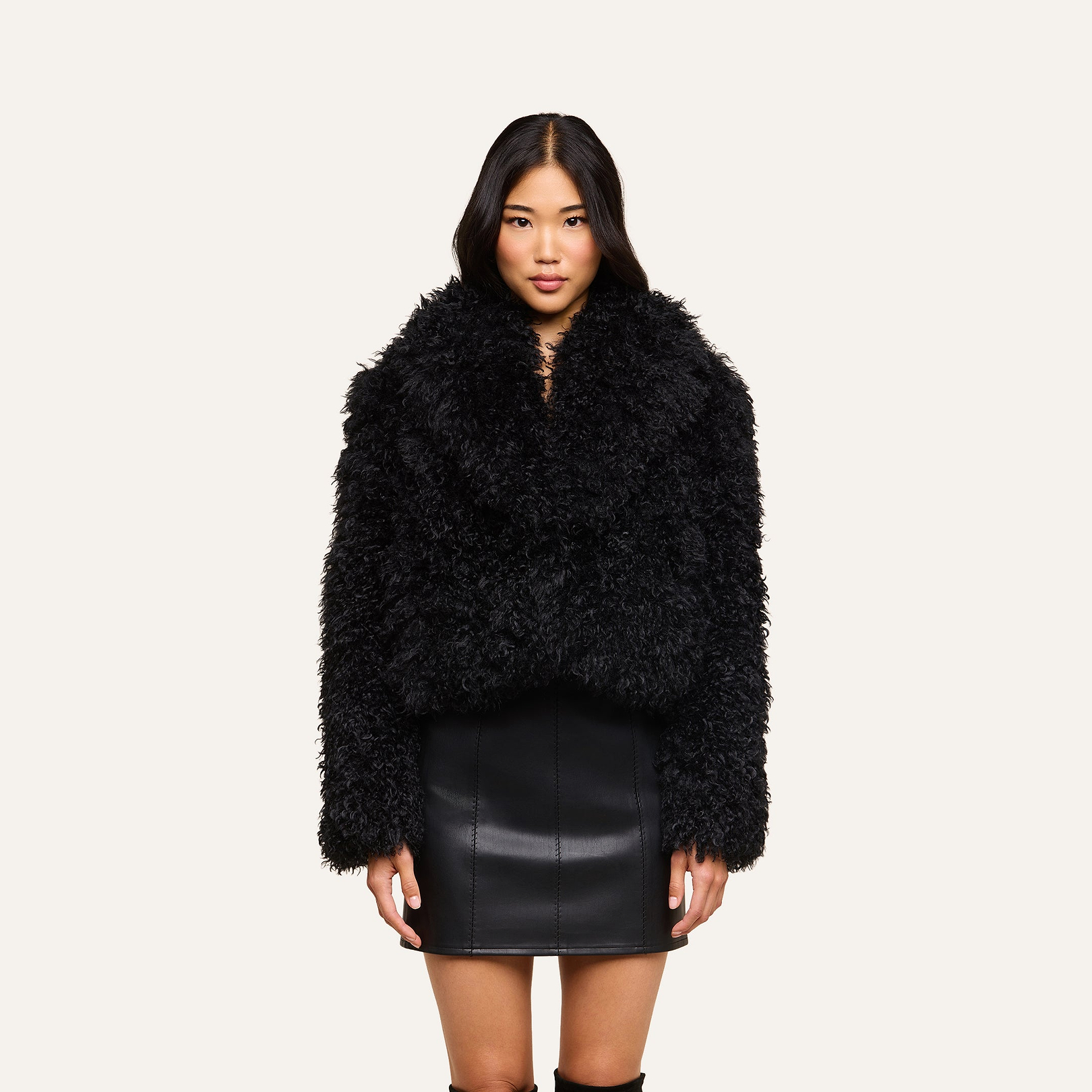 A woman stands against a beige background, wearing the Aura Cropped Faux Fur Jacket as a statement layer over a black leather skirt, looking directly at the camera with her arms relaxed at her sides.