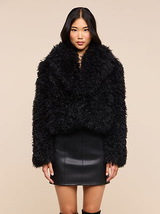 A woman stands against a beige background, wearing the Aura Cropped Faux Fur Jacket as a statement layer over a black leather skirt, looking directly at the camera with her arms relaxed at her sides.