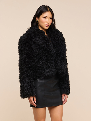 A woman wearing the Aura Cropped Faux Fur Jacket and a black leather skirt stands against a plain beige background, looking slightly to the side with a neutral expression, effortlessly showcasing this bold statement layer.