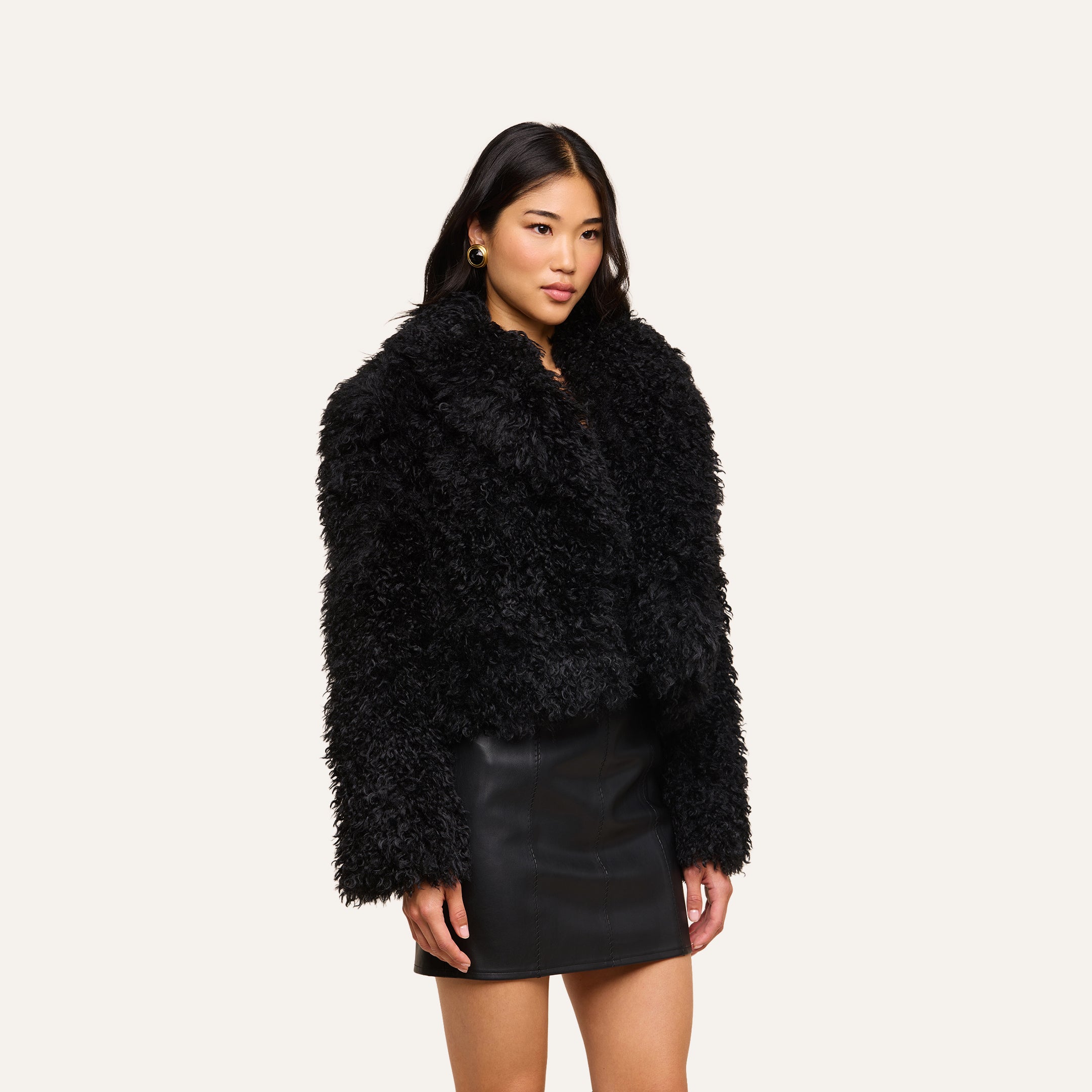Wearing the Aura Cropped Faux Fur Jacket and a black leather skirt, a woman stands against a simple light background, gazing slightly to the side with a neutral expression.