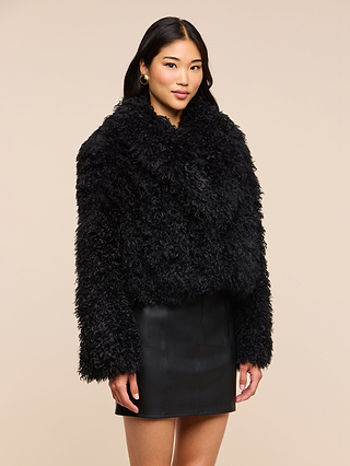 A woman stands against a beige background wearing the Aura Cropped Faux Fur Jacket and a black leather skirt, her hands relaxed at her sides and a slight smile on her face—a perfect statement layer for any modern wardrobe.