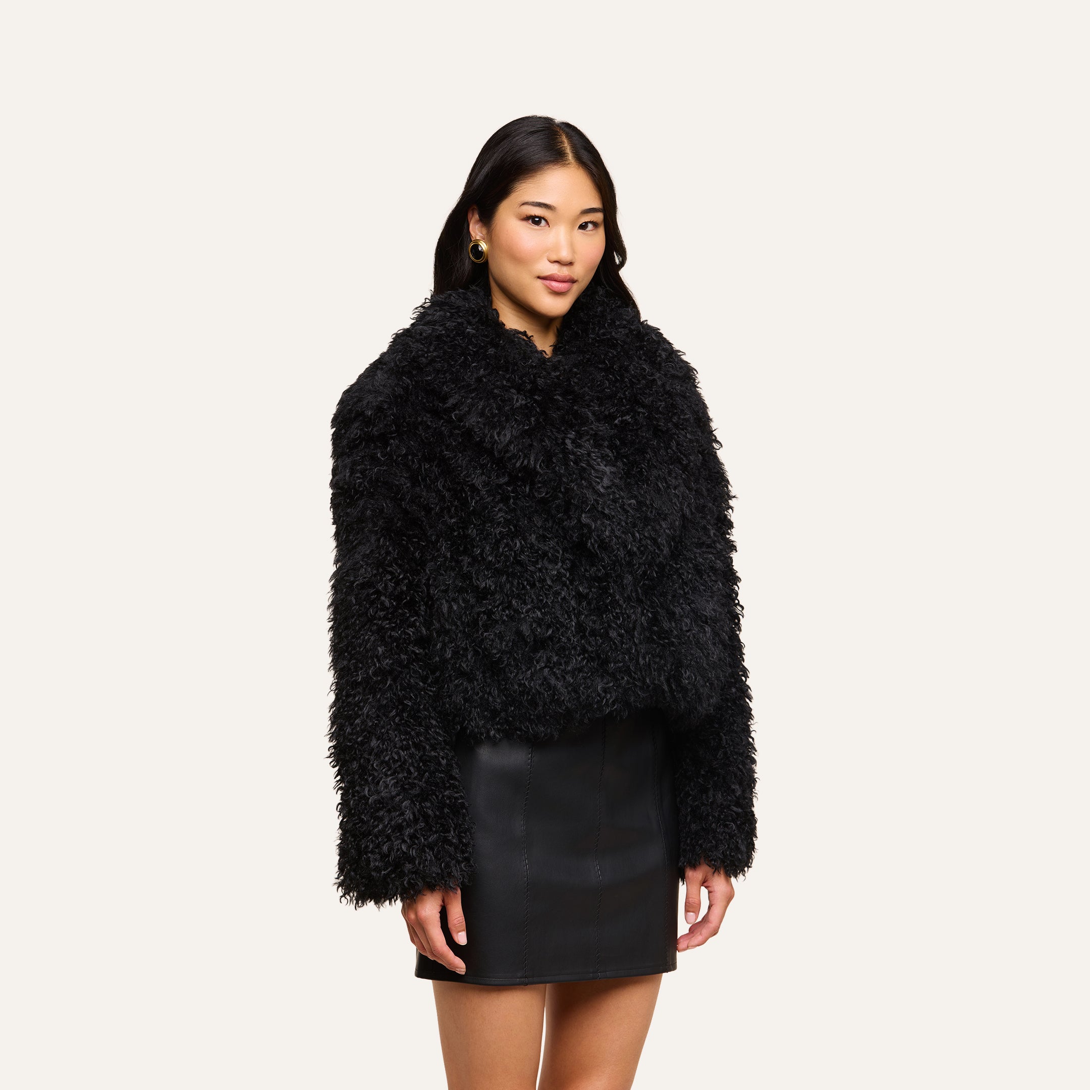 A woman with long dark hair wears the Aura Cropped Faux Fur Jacket and a black leather skirt, standing against a plain light background.