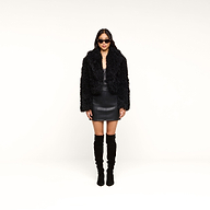 A woman faces forward against a white background, wearing black sunglasses, a black mini skirt, and black knee-high boots. She completes her ensemble with the Aura Cropped Faux Fur Jacket, adding a bold statement layer to her look.