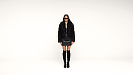 A person wearing sunglasses, a black fuzzy jacket, a black leather skirt, and knee-high black boots stands against a plain white background.