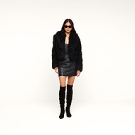 A woman in black sunglasses wears the Aura Cropped Faux Fur Jacket, a black leather mini dress, and black over-the-knee boots, standing against a plain white background.