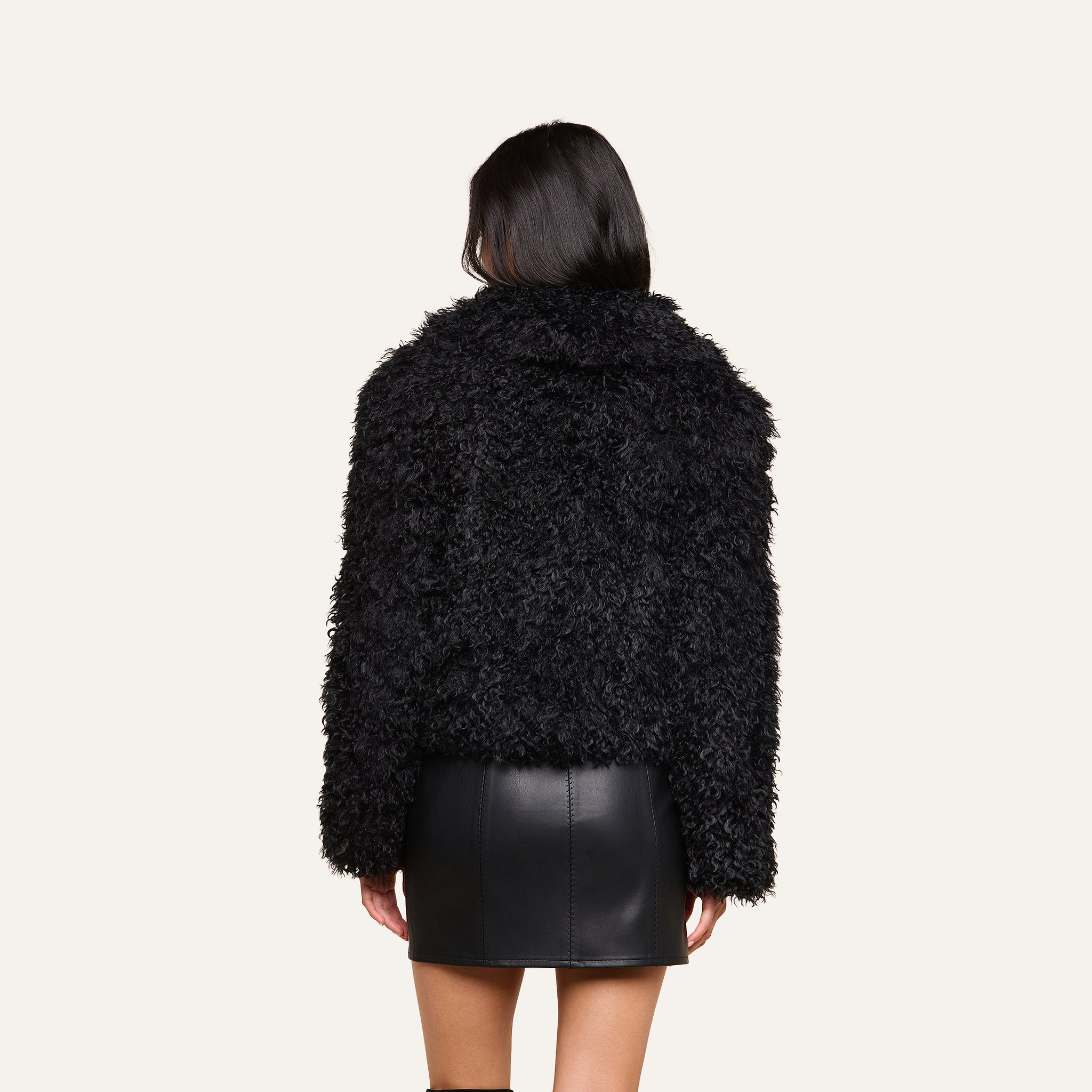 A woman with straight dark hair wears the Aura Cropped Faux Fur Jacket in black along with a black leather mini skirt, standing with her back to the camera against a plain light background.