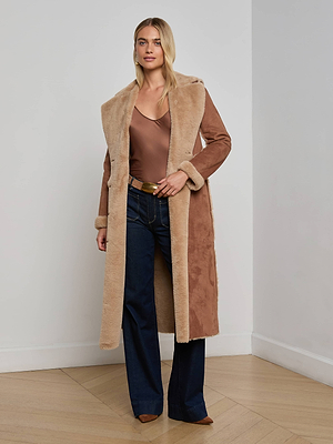 A woman stands indoors wearing the L'AGENCE Citron Faux Fur Maxi Coat over a brown satin top, dark blue flared jeans, and brown pointed shoes, with her hair in loose waves.