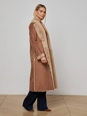 A woman poses indoors against a light gray wall and wooden floor, wearing the L'AGENCE Citron Faux Fur Maxi Coat with beige fur lining, dark blue flared jeans, and brown shoes. The coat’s oversized silhouette offers modern style.