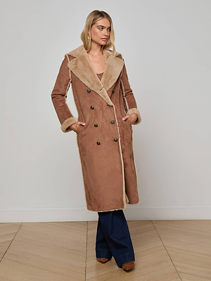 A woman stands indoors wearing the L'AGENCE Citron Faux Fur Maxi Coat with wide lapels, oversized fit, dark buttons, and side pockets. She pairs it with blue flared jeans and brown shoes on a light wood floor against a plain wall.
