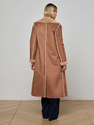 A woman faces away, wearing the L'AGENCE Citron Faux Fur Maxi Coat in brown vegan suede with cream faux shearling trim and cuffs, paired with dark pants. She stands on a light wooden floor against a plain white wall.
