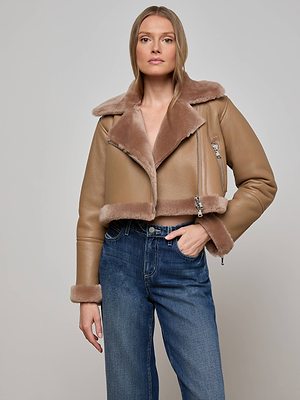 A woman wears the tan Pike Shearling Leather Jacket with a large fur collar and cuffs, styled with blue high-waisted jeans, standing against a plain light gray background.