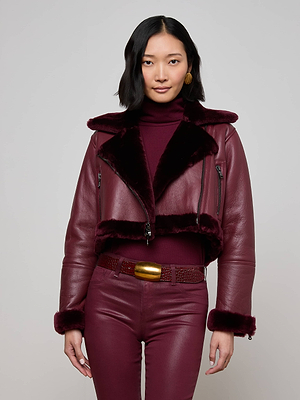 A woman with medium-length black hair models the Pike Shearling Leather Jacket in burgundy with faux fur trim, styled with a matching turtleneck, high-waisted pants, and a gold-buckled braid belt against a plain background.