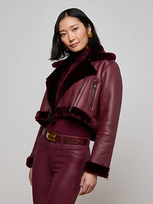 A woman with shoulder-length black hair models the Pike Shearling Leather Jacket—burgundy leather with faux fur trim—styled in a studio with a matching turtleneck, high-waisted pants, gold earrings, and a textured belt.