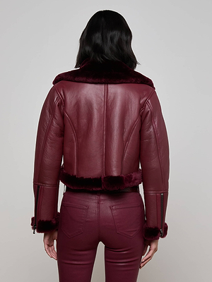A person with shoulder-length dark hair is seen from behind, wearing the Pike Shearling Leather Jacket in burgundy with faux fur details and matching pants, styled as a chic winter layer against a light gray background.