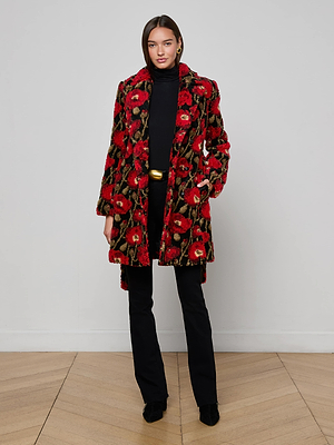 A woman stands indoors against a white wall wearing the L'AGENCE Tristan Sherpa Midi Coat, featuring a bold red poppy print, styled with a black turtleneck, pants, and shoes.