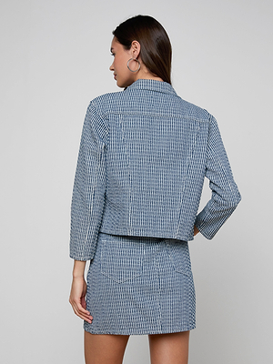 A woman faces away wearing the Nelly Cropped Jacket in blue and white textured cotton with a matching skirt. She has long brown hair, hoop earrings, and rests her left hand on her hip against a plain light background.