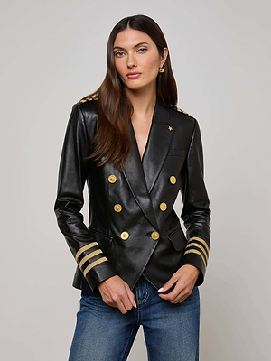 A woman with long brown hair wears the Kenzie Leather Blazer, featuring structured shoulders, gold buttons, striped military-style cuffs, and pairs it with blue jeans while standing against a plain light background.
