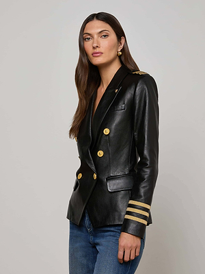 A woman with long brown hair wears the Kenzie Leather Blazer featuring structured shoulders, gold buttons, gold sleeve stripes, and epaulets. She pairs it with blue jeans and stands against a plain light background.