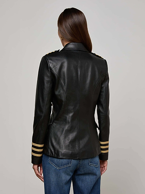 A person with long brown hair, seen from behind, wears the Kenzie Leather Blazer—fitted black leather with gold-striped cuffs and structured shoulders—paired with dark blue jeans, set against a plain neutral background.