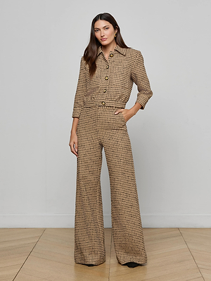 A woman with long brown hair stands by a plain wall in the Jia Cropped Tweed Jacket, featuring brown and beige textured tweed, large buttons, and wide-leg trousers, with one hand in her pocket.
