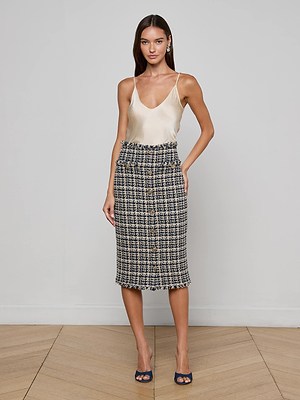 A woman with long brown hair and a neutral expression stands against a plain wall wearing L'AGENCE's Tai Tweed Midi Skirt, a cream satin camisole, and navy open-toe heels.