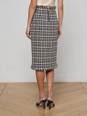 A person wears the Tai Tweed Midi Skirt by L'AGENCE, a high-waisted black and white Italian plaid skirt with subtle gold buttons and a back zipper, paired with navy high-heeled shoes on a wooden floor.